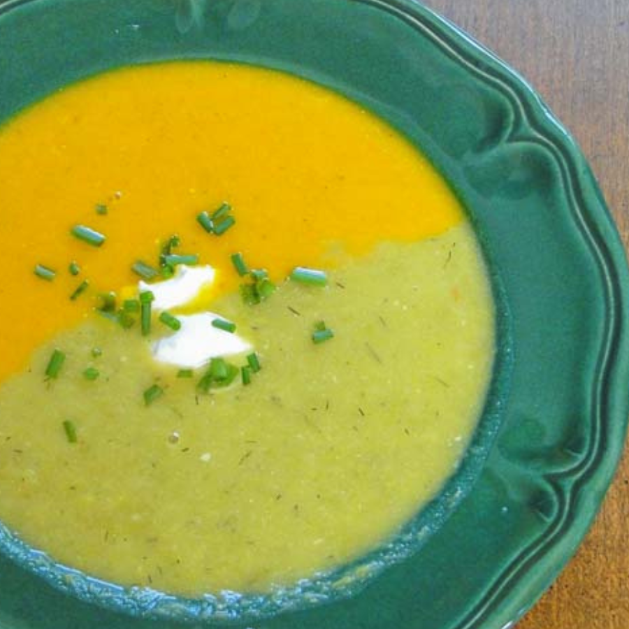 A close-up shot of a green ceramic bowl filled with a split yellow and green soup, garnished with a dollop of sour cream and chopped chives, on a wooden table.