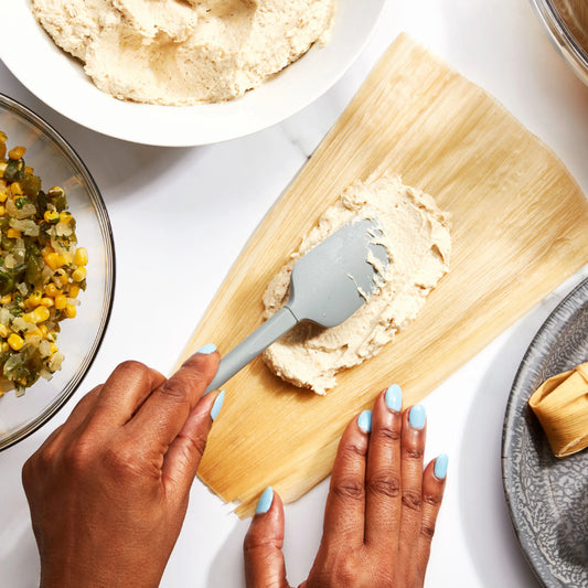 Person spreading a spreadable food item on a corn husk with a spatula, surrounded by ingredients on a white surface.