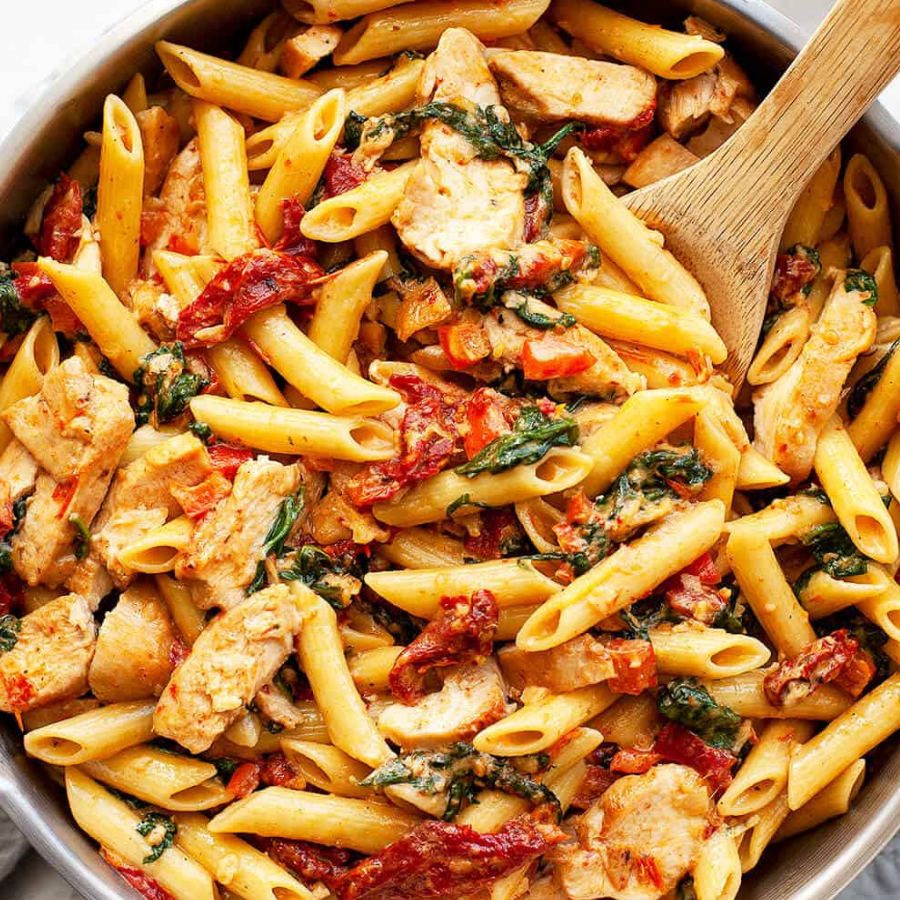 A close-up of a creamy penne pasta dish mixed with sliced chicken, sun-dried tomatoes, and green spinach leaves, served in a metal pan with a wooden spoon.