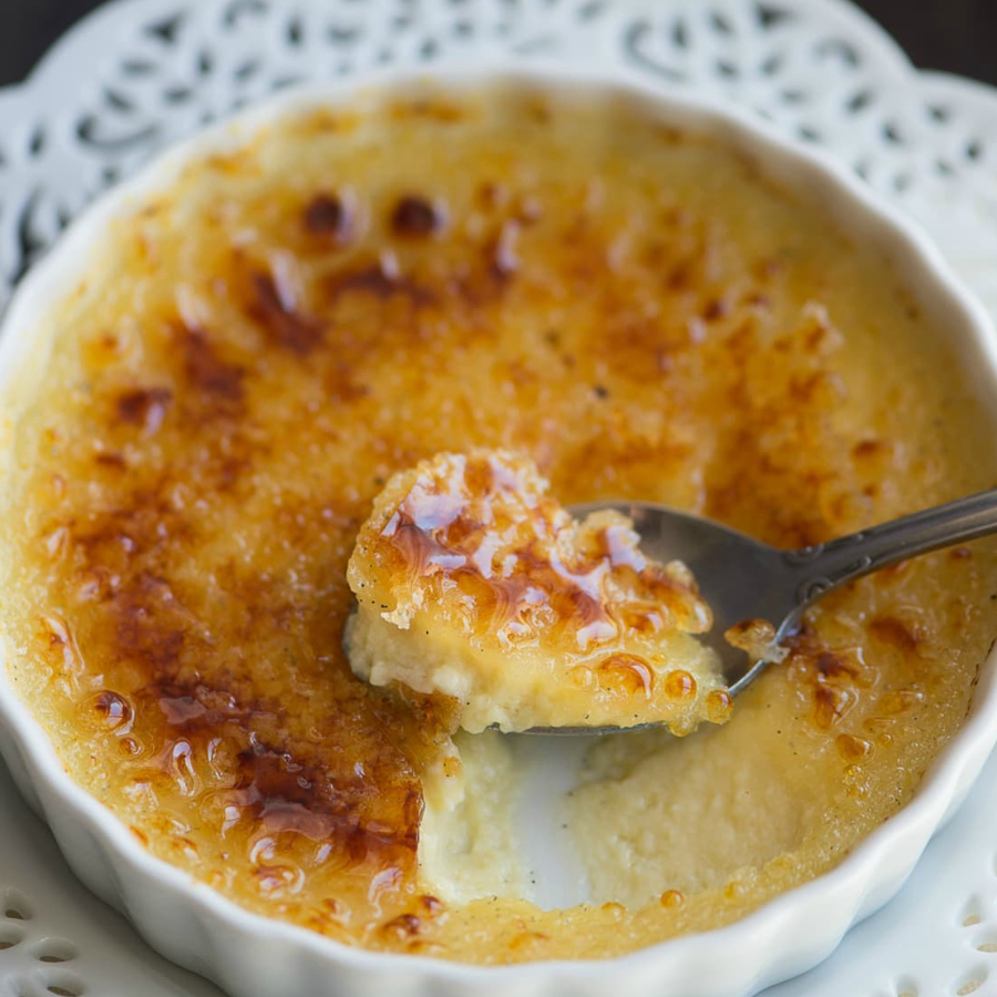 A close-up of a creme brulee dessert in a white ramekin, with a spoon lifting a portion to show the creamy custard underneath and the caramelized top surface.