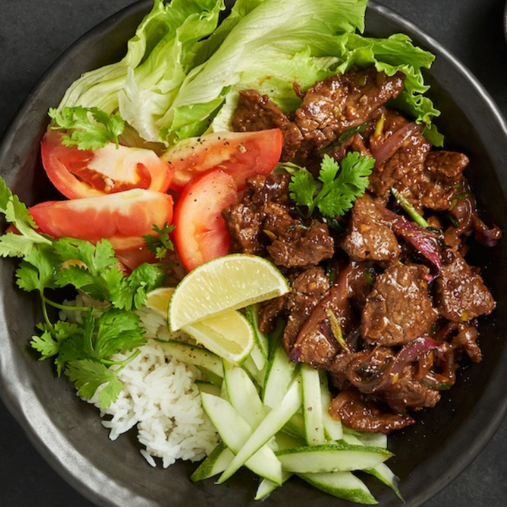 A top-down view of a prepared beef dish served over white rice in a dark bowl, with lettuce, tomato slices, cucumber strips, lime wedges, and cilantro on a dark tabletop.