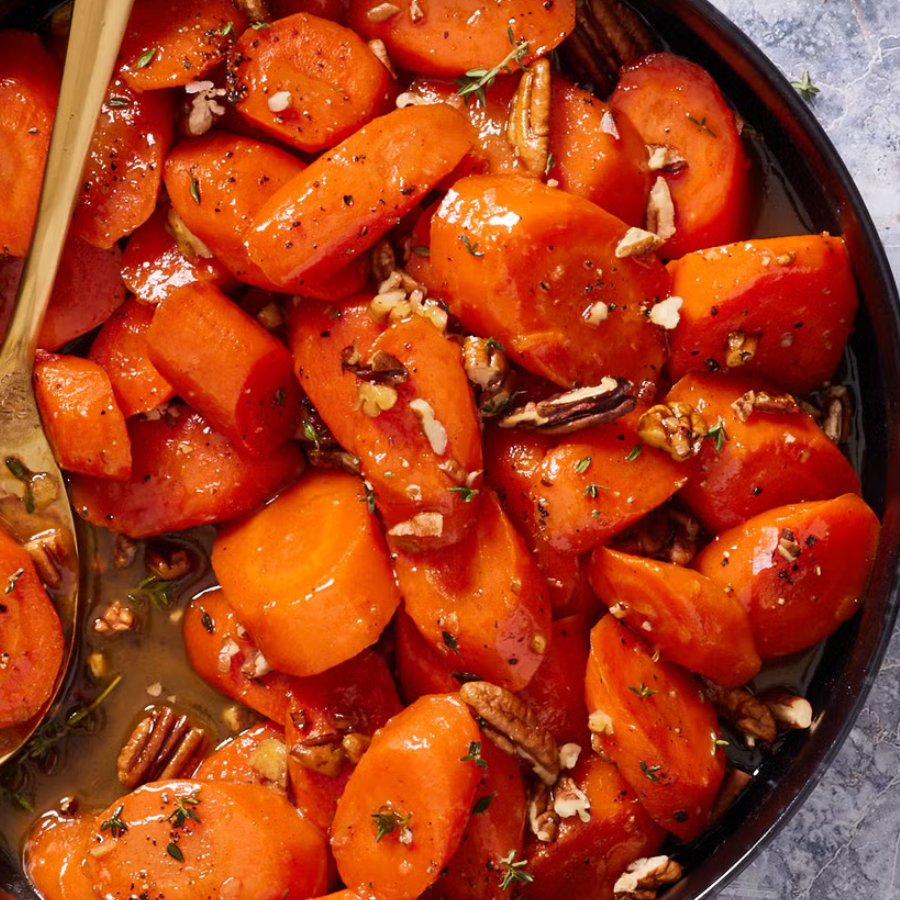 Close-up overhead of a cooked dish of orange carrots glazed with sauce, topped with chopped pecans and herbs, served in a dark round pan with a gold spoon visible on the left.
