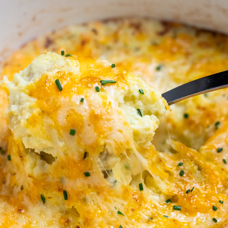 Close-up of cheesy baked potato or mashed potato with melted cheddar cheese, chives, and a spoon lifting a portion.