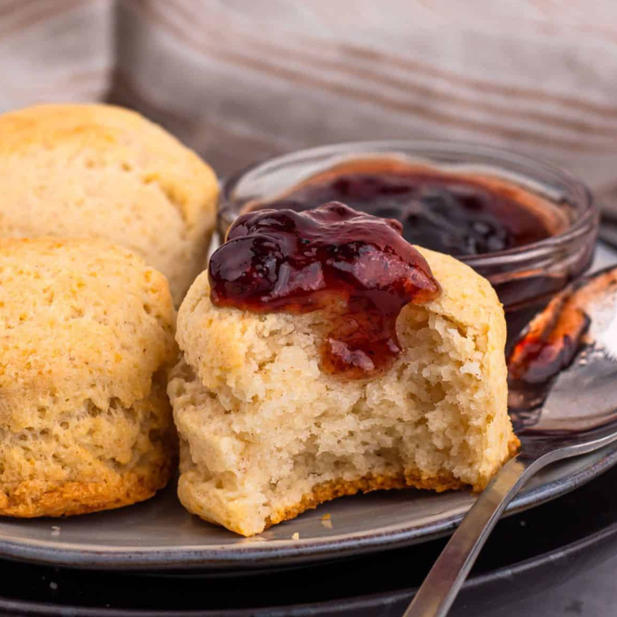 A close-up of golden-brown scones on a plate, with one cut open and topped with dark berry jam, alongside a small glass bowl of jam and a spoon.