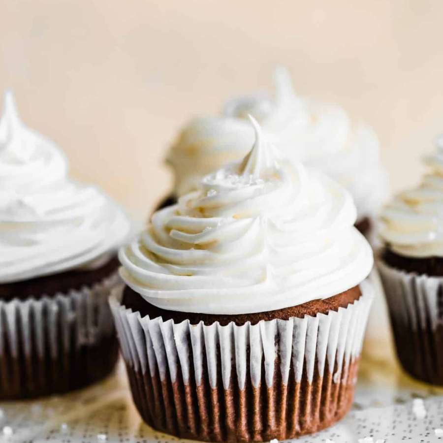 A close-up of a chocolate cupcake with thick white whipped frosting and a sprinkle of coarse salt on top, set against a softly blurred warm beige background.