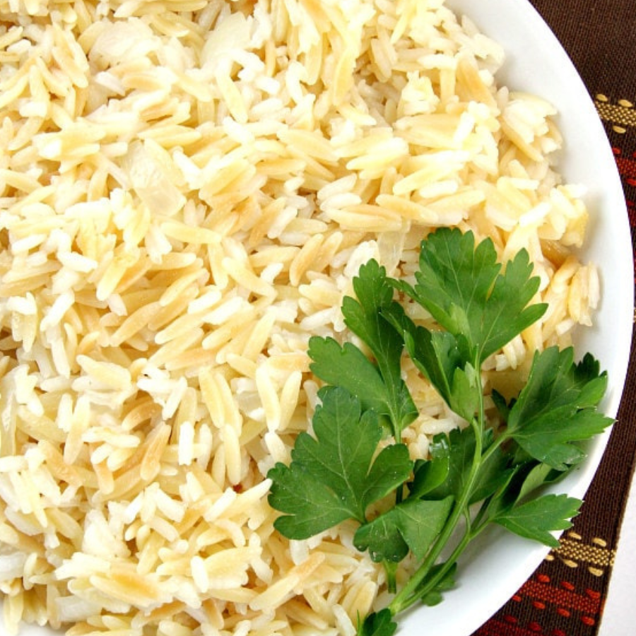 A close-up of cooked rice on a white plate, garnished with a sprig of fresh green parsley, with a patterned fabric visible at the edge.