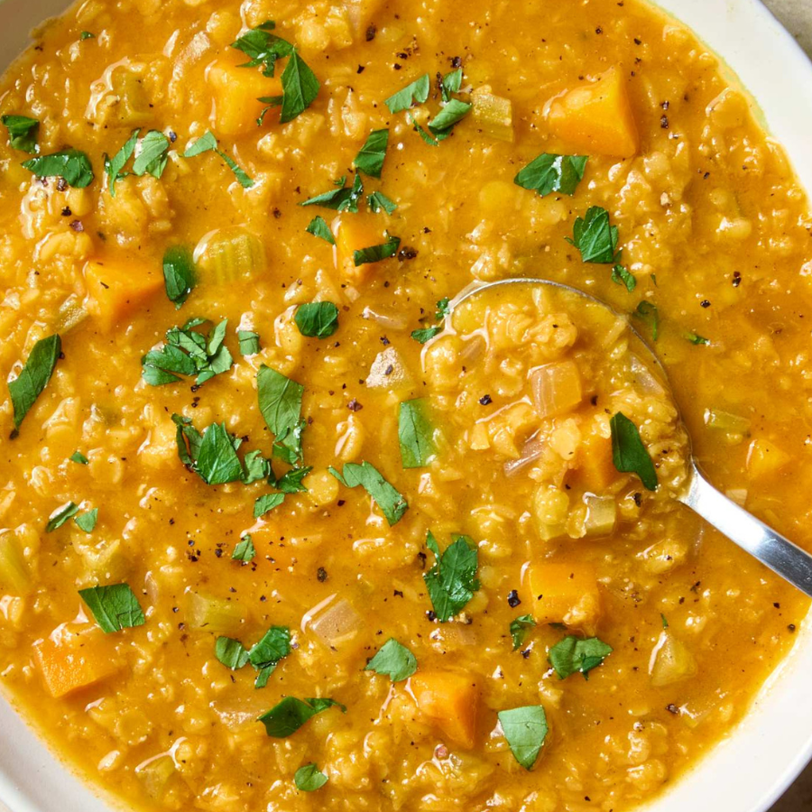 A close-up overhead shot of a bowl of chunky lentil soup with butternut squash, celery, and fresh green parsley garnish, with a metal spoon resting in the bowl.