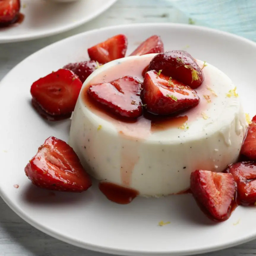 A white molded dessert, likely a cheese cake or custard, topped with sliced glossy strawberries and a drizzle of syrup, served on a white plate.