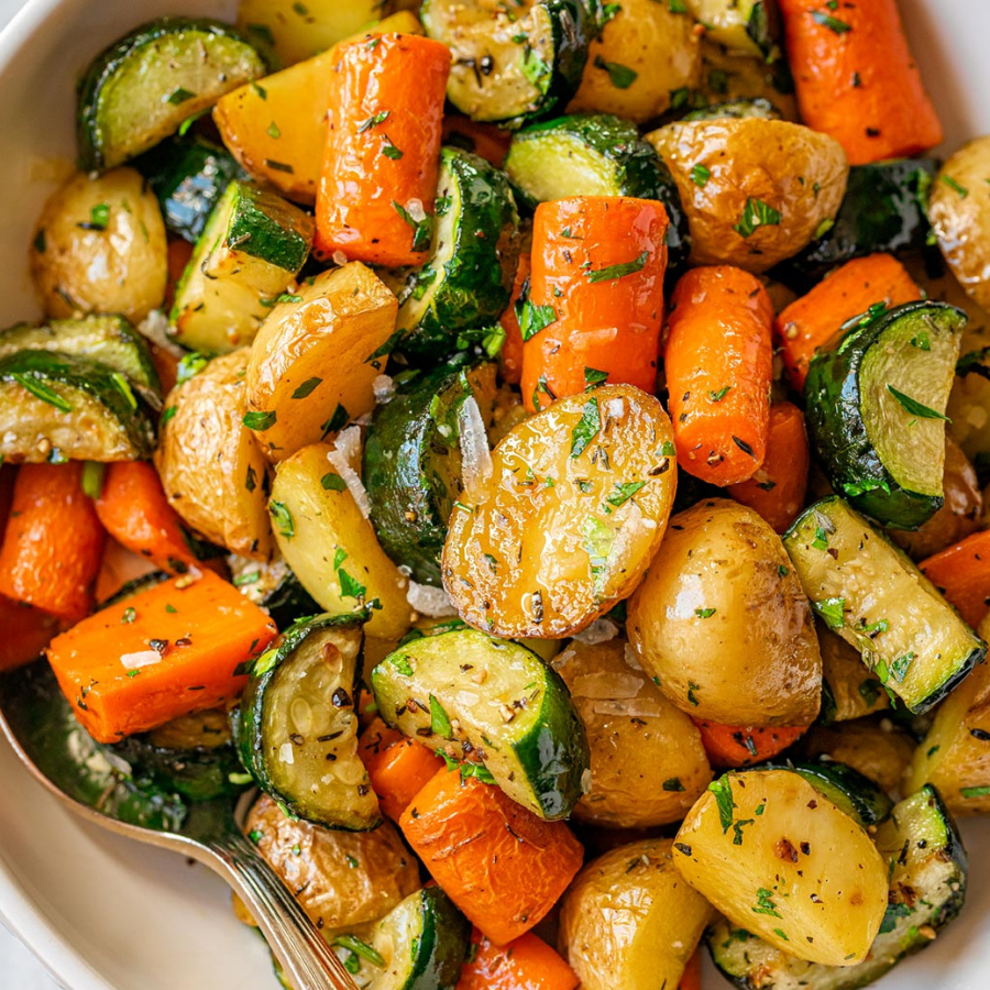 A close-up overhead view of a mixed vegetable medley in a white bowl, including roasted potatoes, carrots, zucchini, and onions with a light sauce and herbs.