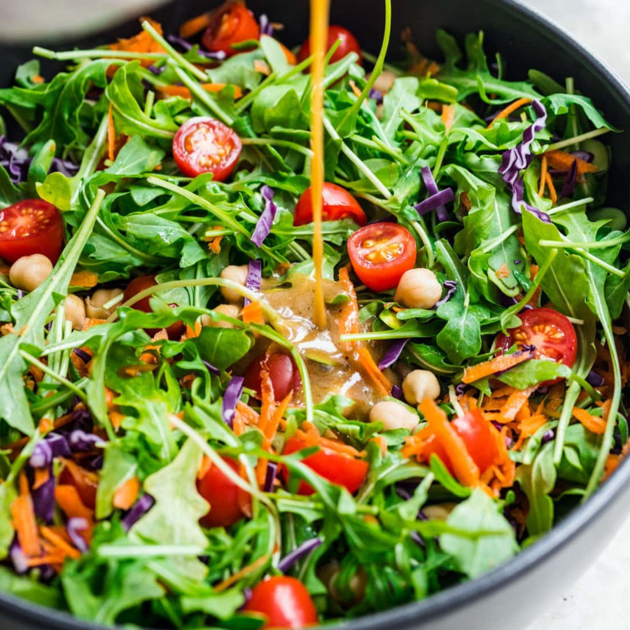 A close-up of a fresh green salad in a black bowl, featuring arugula, cherry tomatoes, shredded carrots, purple cabbage, chickpeas, and a drizzle of dressing being poured from above.