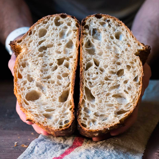 A close-up of a person holding a freshly baked sourdough bread loaf, sliced vertically in half to show the airy, porous crumb structure. The hands and part of the torso are visible, with a rustic wooden table and a cloth towel in the background.