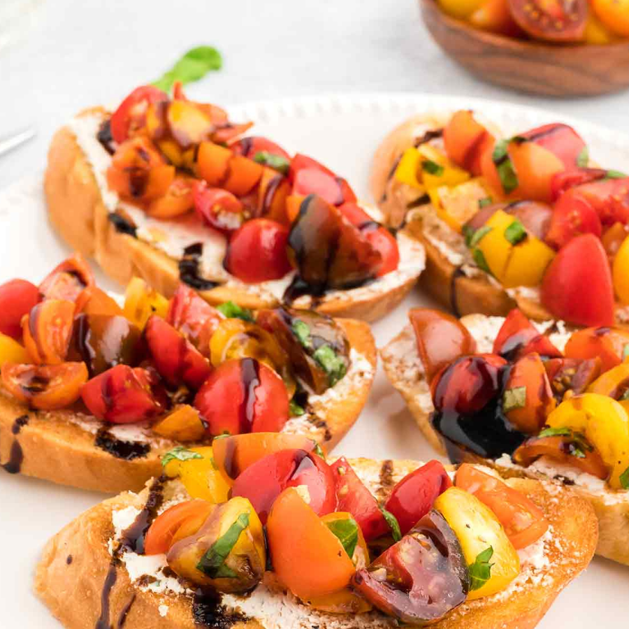 Close-up of several slices of toasted bread topped with white cheese, roasted tomatoes, yellow peppers, and a drizzle of balsamic glaze on a white plate.