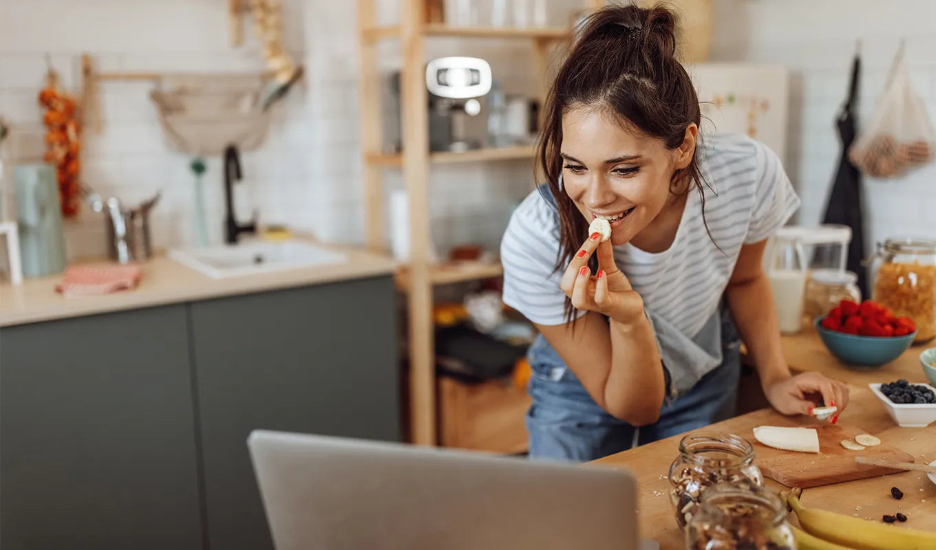 Woman in a kitchen eating a piece of pizza while looking at a laptop.